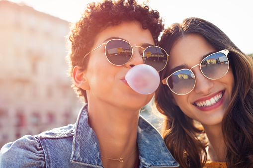 Portrait of brazilian woman with curly hair making bubble while latin girl smiling. Multiethnic friends enjoying vacation in city and looking at camera. Lesbian couple having fun while inflating chewing gum and making pink balloon.