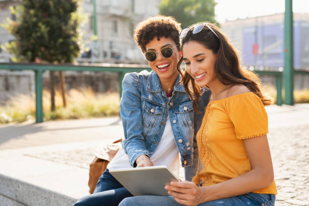 две женщины, использующие цифровой планшет на открытом воздухе - city cheerful urban scene happiness стоковые фото и изображения