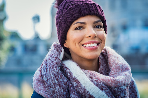 Closeup face of a young happy african woman enjoying winter wearing scarf and cap. Smiling brazilian girl in a colorful shawl looking at camera in a cold day. Multiethnic woman with knitted bordeaux hat and woolen scarf.