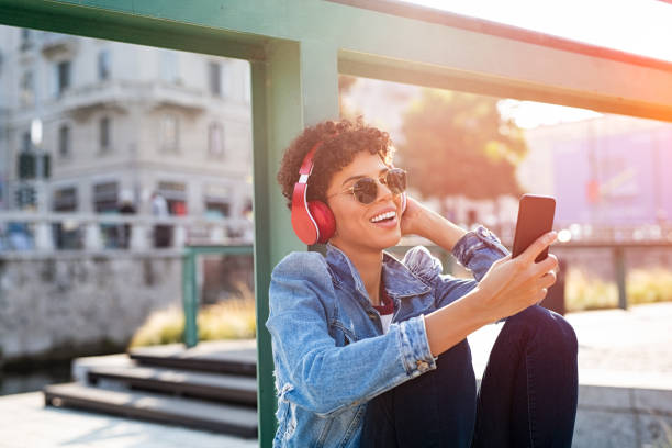 Young woman having fun with music Young beautiful woman holding smartphone and listening to music with wireless headphones. Cheerful smiling girl with curly hair and red headphones changing music track on phone. Carefree urban teen enjoying songs while sitting on ground. double denim stock pictures, royalty-free photos & images