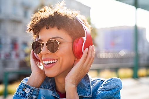 Young smiling african woman relaxing and listening music with headphones at sunset. Brazilian smiling girl listening songs via wireless headphones on the street. Closeup face of teen wearing sunglasses and keeps the rhythm of the song.