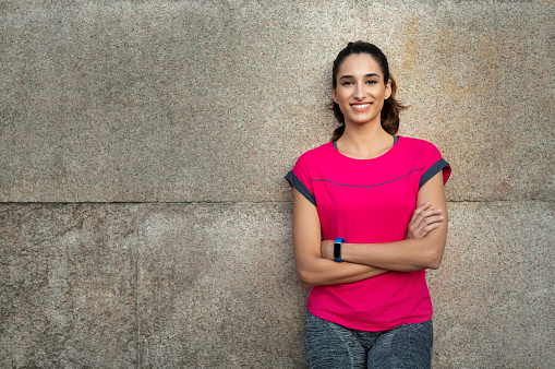 Smiling sporty young woman take a break after running. Confident latin girl leaning against wall with arms crossed in sportswear. Portrait of happy runner looking at camera and smiling with crossed arms, copy space.
