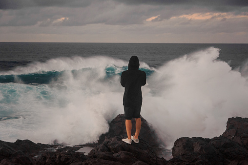 Young man in hoody and shorts standing on the rock near the stormy sea with high waves with foam under the grey sky
