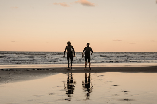 Two physically fit young men walking in black wetsuits with boards walking into the sea in twilight