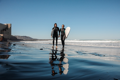 Young couple of surfers walking and laughing along the sea shore with black sand on sunny day