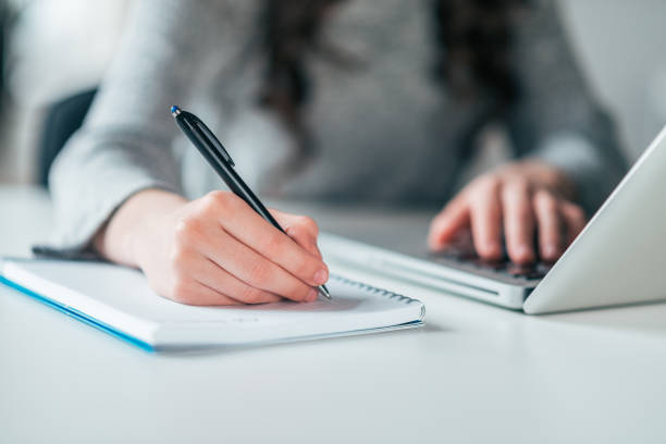 istock Front view of female student writing in notebook and using laptop, close-up. 1134018465
