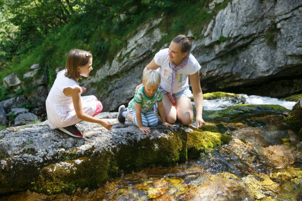 Mother with kids talking, drinking water from a pure, fresh and cool mountain stream on a family trip. Outdoor lifestyle, natural parenting, childhood experience concept.