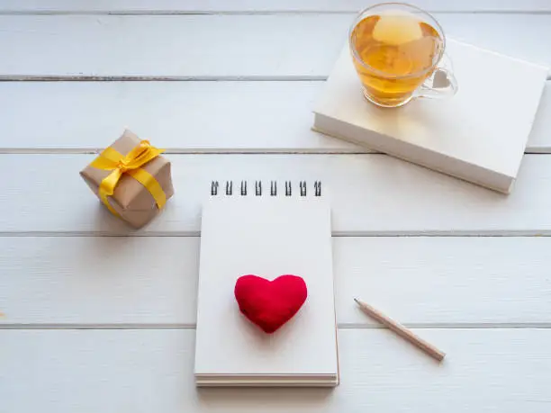 Notepad with a red heart, a cup of tea and a gift on a white wooden table.