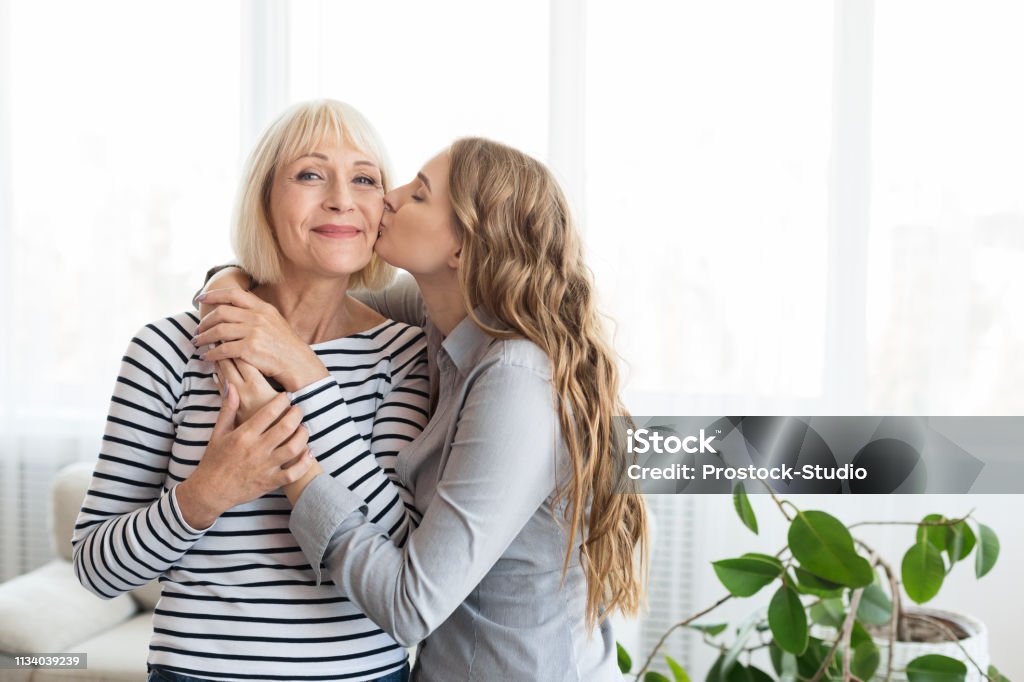 Woman kissing senior mother on the cheek Woman kissing senior mother on the cheek, standing near window at home Mother Stock Photo