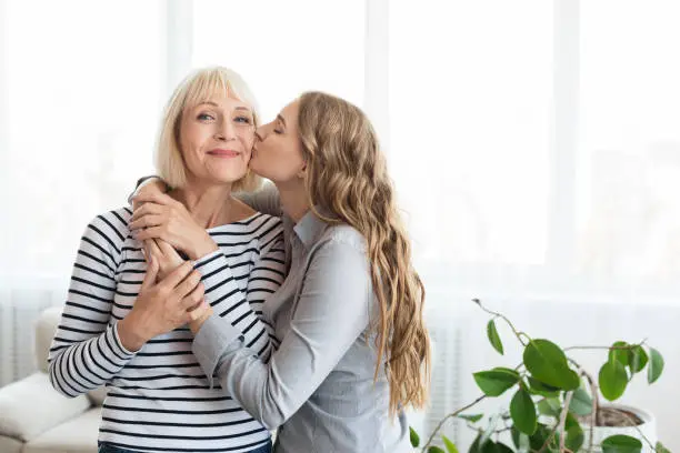 Woman kissing senior mother on the cheek, standing near window at home