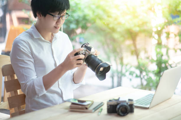 istock Young Asian man holding digital single-lens reflex camera (DSLR) checking photo on camera screen display sitting with laptop computer and smartphone on the table. Photography, art and hobby concepts 1134039282