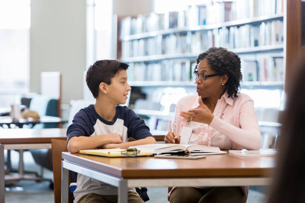 Middle school age boy receives homework help from mentor A middle school age boy sits at a table in a school library with his mature female mentor.  He listens as she gestures and speaks. teacher classroom child education stock pictures, royalty-free photos & images