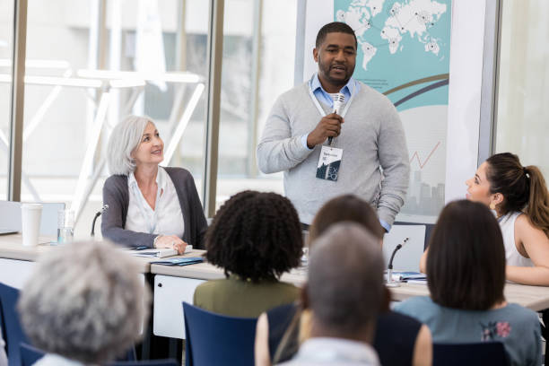 Confident mid adult businessman speaks during conference Mid adult African American man holds a microphone while addressing an audience during a panel discussion. summit meeting photos stock pictures, royalty-free photos & images
