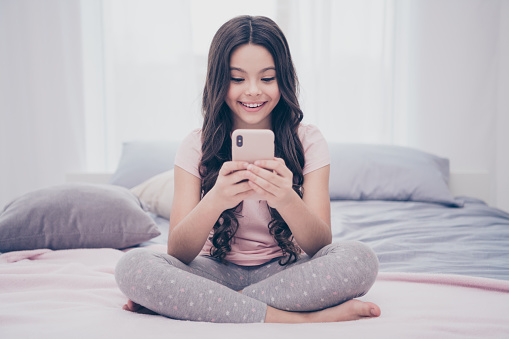 Close up photo cute beautiful she her little girl sitting big bed linen homey sunday smart phone hands arms video call wear home t-shirt pants comfortable apartments flat bright light colored room.