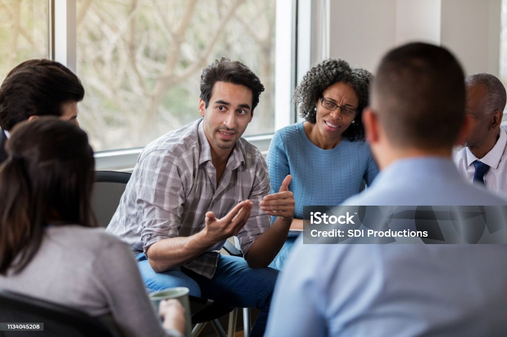 Mid adult businessman gestures during meeting Mid adult businessman discusses something with a group of colleagues. He gestures while talking. Group Of People Stock Photo