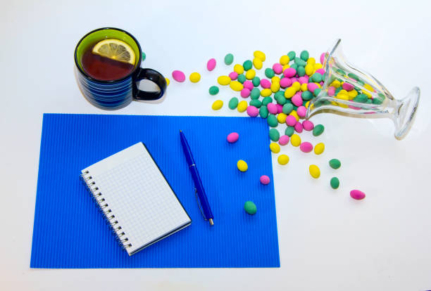 Blank spiral notepad, pen, cup of tea with lemon slice and glass vase with colorful candies on the table.