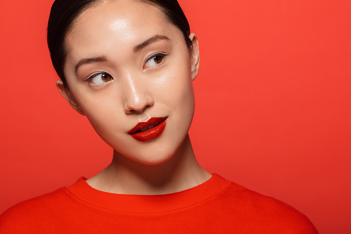Close up of young asian woman with beautiful make up looking away and thinking. Korean female model with red make up against red background.