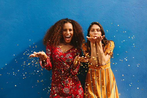 Female friends blowing off magic glitter. Excited women having fun against blue wall.