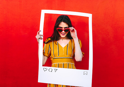Woman wearing sunglasses holding a empty social media photo frame. Stylish female content creator standing against red looking through at empty photo frame.