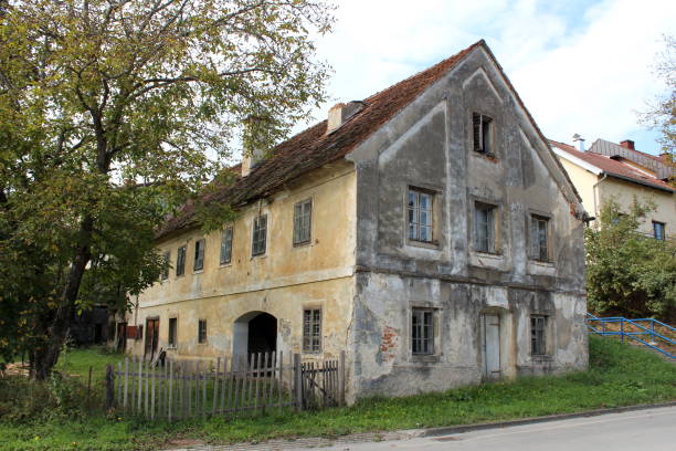 istock Large old family house with multiple windows and dilapidated cracked facade surrounded with uncut grass and tall trees next to paved road 1134078188