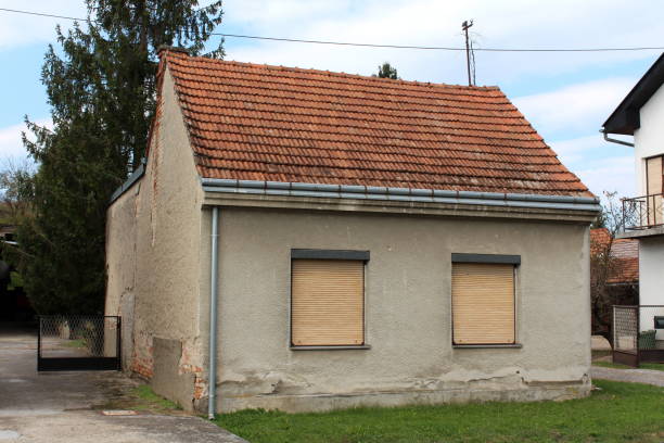 istock Small abandoned family house with dilapidated facade and two front windows covered with window blinds surrounded with grass and other houses 1134078240