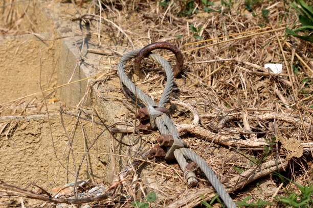 istock Strong metal cable connected to rusted ring built into concrete foundation and held in place with screws and bolts surrounded with dry grass 1134078243