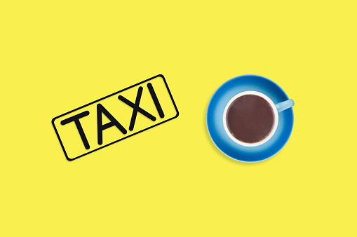 Word taxi in frame of black color cut out of black paper near full blue cup of hot chocolate on yellow background. Top view. Concept of journey. Copy space