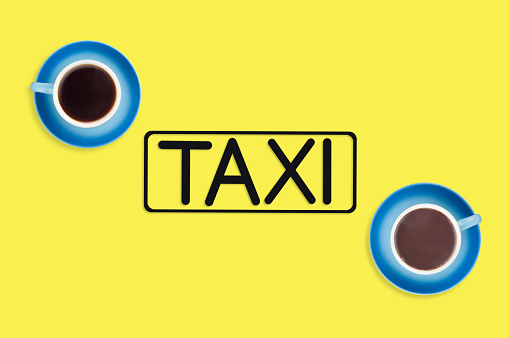 Word taxi in frame of black color cut out of black paper near full blue cups of hot chocolate and coffee on yellow background. Top view. Concept of journey