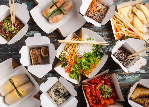 Assorted Chinese dishes in paper delivery boxes: sweet and sour chicken, dim sum, spring rolls, noodles, salad, rice, steamed buns, dips. Asian restaurant take away concept, top view