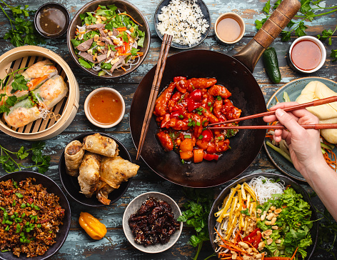 Set of assorted Chinese food on table with female hand holding chopsticks from above. Full and festive table with all traditional Chinese dishes, asian style dinner or buffet, top view