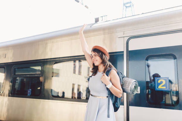 テーマの輸送と旅行。歯笑顔を持つ若い白人女性の肖像電車の駅の背景に立っているバックパックと手を振ってこんにちは、夏のドレスと帽子で喜びのサイン - waving separation bye women ストックフォトと画像