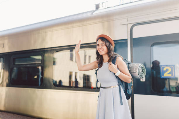 テーマの輸送と旅行。歯笑顔を持つ若い白人女性の肖像電車の駅の背景に立っているバックパックと手を振ってこんにちは、夏のドレスと帽子で喜びのサイン - waving separation bye women ストックフォトと画像