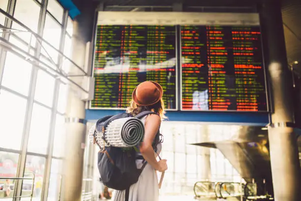 Theme travel public transport. young woman standing with back in dress and hat behind backpack and camping equipment for sleeping, insulating mat looks schedule on scoreboard airport station sunny day