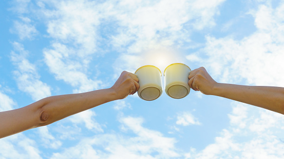 Two Asian woman hands clink hot coffee mug outdoor in the morning. Friends enjoy drinking coffee together. Clear sky background.