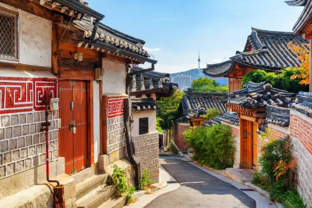 Awesome view of cozy old narrow street and traditional Korean houses of Bukchon Hanok Village in Seoul, South Korea. Seoul Tower on Namsan Mountain is visible on blue sky background. Scenic cityscape.