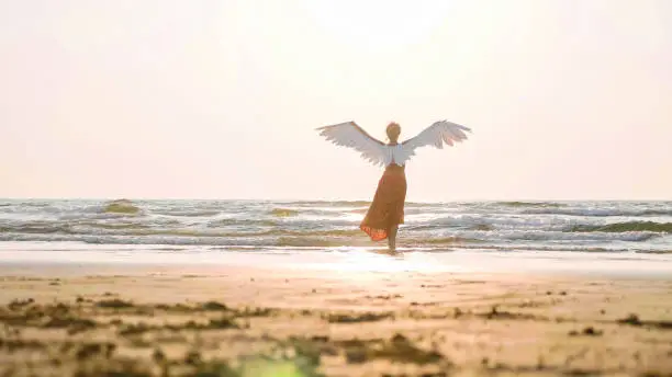 Beautiful female angel walking barefoot from camera toward the sea at sunset, rear view