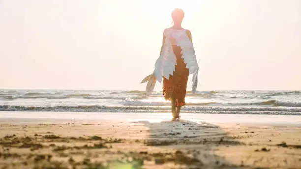 Beautiful female angel walking barefoot from camera toward the sea at sunset, rear view