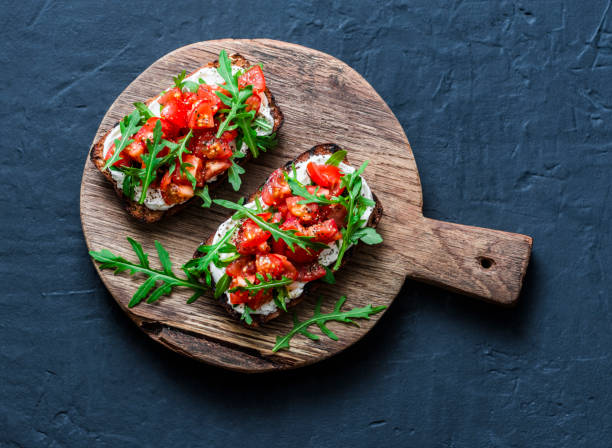 Tomatoes and rocket salad whole grain bread bruschetta on a wooden chopping board on a dark background, top view. Copy space Tomatoes and rocket salad whole grain bread bruschetta on a wooden chopping board on a dark background, top view. Copy space crostini stock pictures, royalty-free photos & images