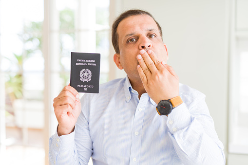 Middle age man holding holding passport of Italy cover mouth with hand shocked with shame for mistake, expression of fear, scared in silence, secret concept