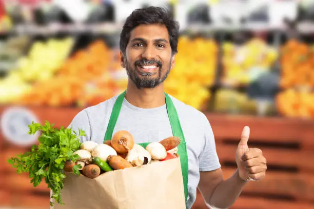 Friendly indian male supermarket or hypermarket employee holding brown paper grocery bag with thumb up as like gesture