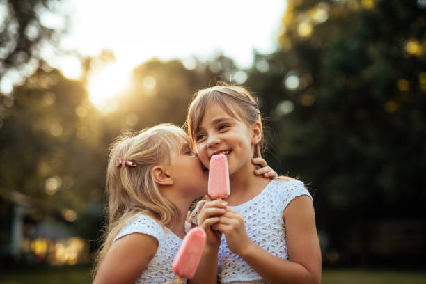 Enjoying nature outdoors Shot of a two young girls whispering while eating ice cream outdoors at sunset. frozen sweet food photos stock pictures, royalty-free photos & images