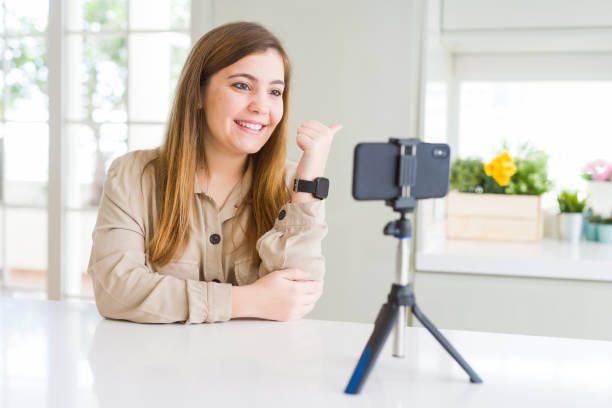 istock Beautiful young woman doing online video call using smartphone webcam smiling with happy face looking and pointing to the side with thumb up. 1134223204