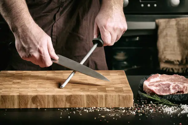 Closeup of male chef hands sharpen a big chef's knife to cut a piece of raw meat on a cutting board.