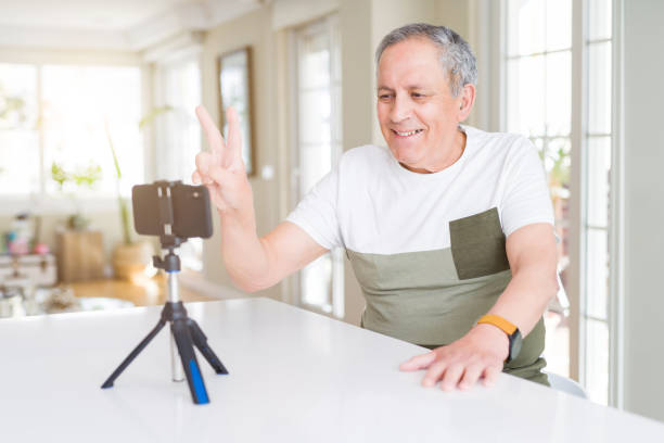 istock Handsome senior man doing video conference talking to the smartphone camera smiling with happy face winking at the camera doing victory sign. Number two. 1134236917