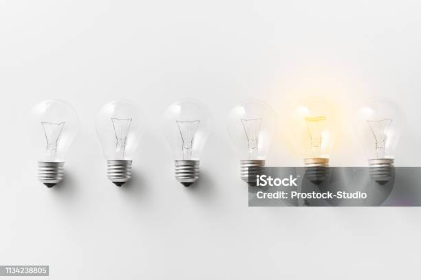 Illuminated Light Bulb In Row Of Dim Ones Stock Photo - Download Image Now - Light Bulb, White Background, Standing Out From The Crowd
