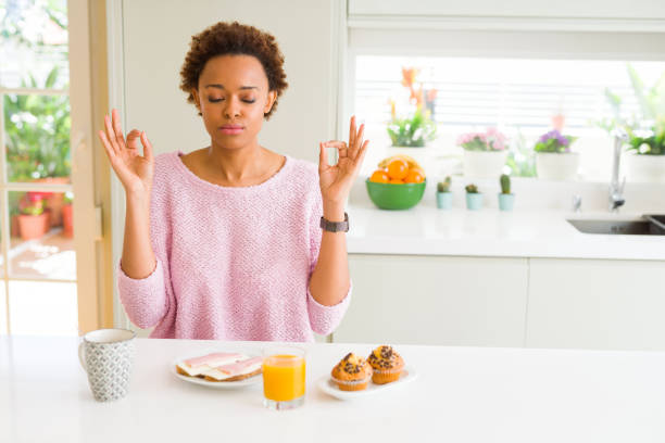 giovane donna afroamericana che mangia breakfast al mattino a casa rilassarsi e sorridere con gli occhi chiusi facendo gesto di meditazione con le dita. concetto di yoga. - african descent american culture exercising women foto e immagini stock
