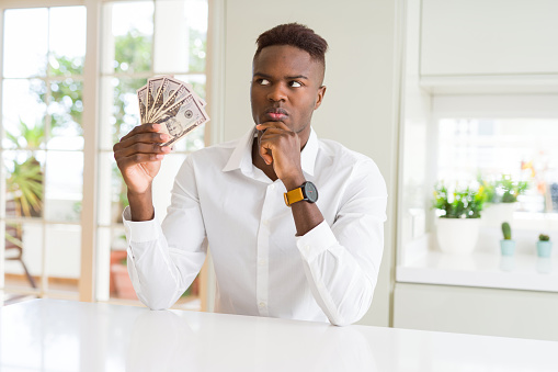 African american man holding fifty dollars bank notes serious face thinking about question, very confused idea
