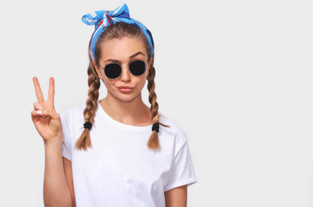 retrato do estúdio da mulher nova loura alegre que desgasta óculos de sol na moda, t-shirt branco e headband azul, fazendo uma face do pato e mostrando o sinal de paz. menina do estudante que vai louca com penteado das tranças - t shirt child white portrait - fotografias e filmes do acervo