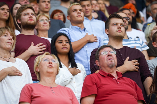 People on a stadium with hands on their hearts listening to and singing the national anthem.