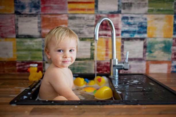 istock Cute smiling baby taking bath in kitchen sink. Child playing with foam and soap bubbles in sunny kitchen with rubber ducks 1134253402